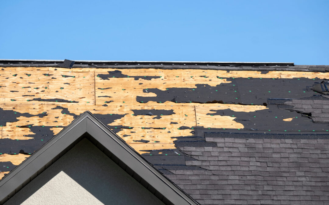 A house roof with missing and damaged shingles, exposing the wooden underlayment beneath—a clear sign of roof damage. Blue sky is visible in the background, highlighting the need to file a claim for proper insurance coverage.